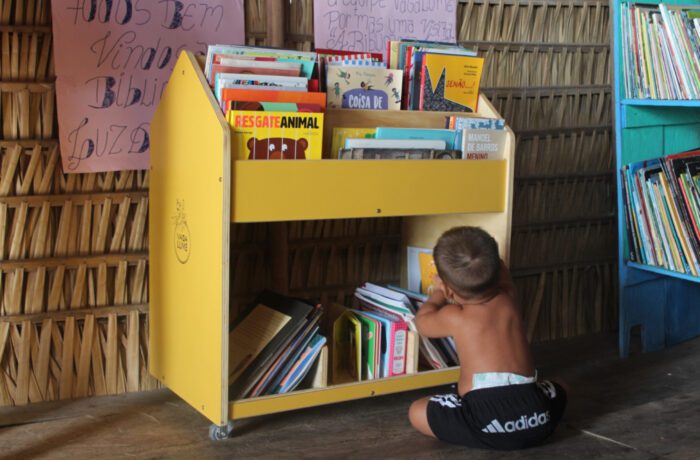 criança sentada no chão pegando livros no expositor de madeira  pintado de amarelo com vários livros infantis