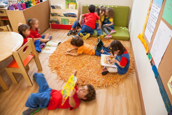 sala de aula da Beacon School com crianças sentadas e deitadas em tapetes no chão lendo livros, a esquerda moveis infantis de marcenaria