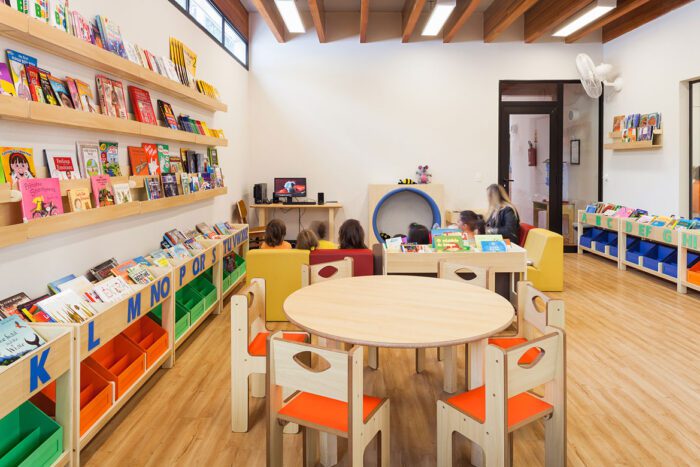 sala de aula com cadeiras e mesas de marcenarias, estantes com livros e ao fundo crianças reuniadas em circulo sentadas em estofado