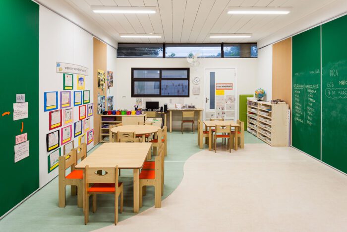 sala de aula com cadeiras e mesas de marcenarias, estantes com livros e ao fundo crianças reuniadas em circulo sentadas em estofado