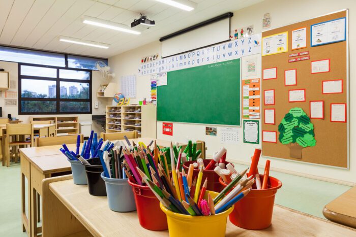 sala de aula com potes de lápis e canetas em cima de mesa, ao fundo quadro negro e painel de avisos