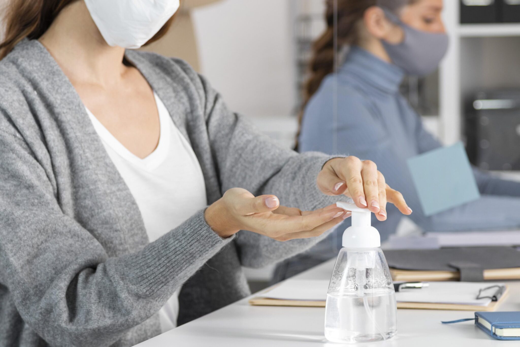 foto aproximada de mulher sentada em sua estação de trabalho higienizando as mãos com frasco de álcool em gel