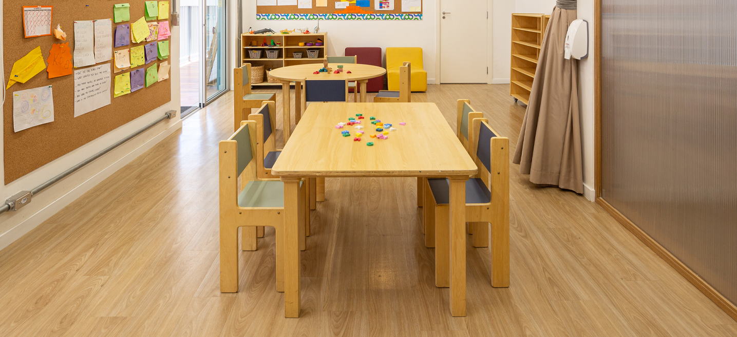 sala de aula infantil com mesas coletivas e cadeiras de madeira, na esquerda painel de recados e ao fundo estantes baixas